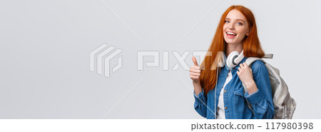 Waist-up portrait joyful cute redhead girl inviting freshmen apply univeristy, got scholarship, smiling showing thumbs-up in approval, recommendation, holding backpack and headphones 117980398