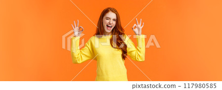 Cheerful and positive, enthusiastic redhead female in yellow sweater winking in approval, smiling joyful, give permission, positive reply, rate good movie, nod agreement, recommend, orange background 117980585