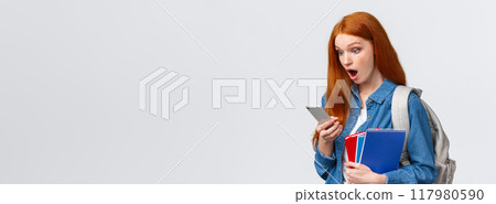 Awesome no classes today. Amazed and excited redhead teenage girl gasping as reading cool message on smartphone, holding backpack notebooks and mobile phone, white background 117980590