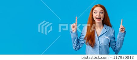 Excited cute redhead girl in nightwear smiling amused and fascinated, telling about incredible choices, great opportunity, pointing fingers up staring camera dreamy and amazed, blue background Excited cute redhead girl in nightwear smiling amused and fascinated, telling about incredible choices, great opportunity, pointing fingers up staring camera dreamy and amazed, blue background 117980818