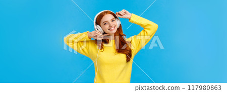 Technology, e-shopping and people concept. Modern good-looking redhead female in yellow sweater, dancing carefree, listen favorite singer music in headphones, smiling camera, blue background 117980863