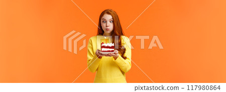 Funny and silly cute redhead b-day girl squinting eyes making goofy expression, pouting as holding birthday cake and blowing-out candle to make wish, having fun at party celebrating, orange wall 117980864