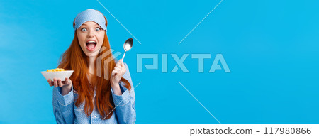 Excited, overwhelmed happy redhead caucasian woman eating cereals morning, holding spoon, smiling amused and amazed staring camera, wear sleep mask, pyjama, standing blue background Excited, overwhelmed happy redhead caucasian woman eating cereals morning, holding spoon, smiling amused and amazed staring camera, wear sleep mask, pyjama, standing blue background 117980866