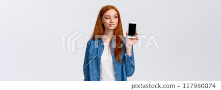 Curious and daring sassy redhead girl tempting buy something in online store, have interesting plan, smirk and peek at smartphone screen with pleased expression, standing intrigued white background 117980874
