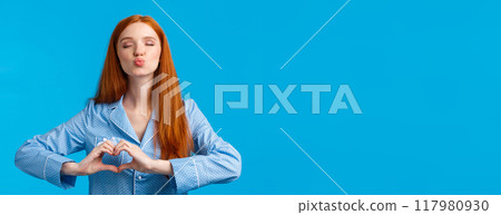 Kiss for goodnight. Charming pretty redhead female in pyjama folding lips and close eyes give mwah, showing heart gesture over chest, express affection or tender feelings, blue background Kiss for goodnight. Charming pretty redhead female in pyjama folding lips and close eyes give mwah, showing heart gesture over chest, express affection or tender feelings, blue background 117980930