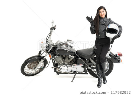 A biker girl standing next to her bike, holding a helmet and pointing A biker girl standing next to her bike, holding a helmet and pointing 117982932