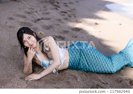 年輕的女美人魚躺在沙灘的樹蔭下微笑 117982940