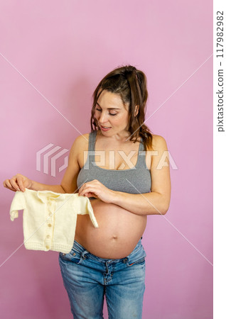 Expecting mother in a cozy home setting, tenderly holding up a knitted sweater 117982982