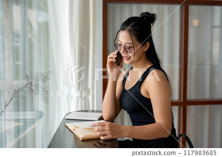 A beautiful Asian woman is looking for information in a book while talking on the phone. A beautiful Asian woman is looking for information in a book while talking on the phone. 117983685
