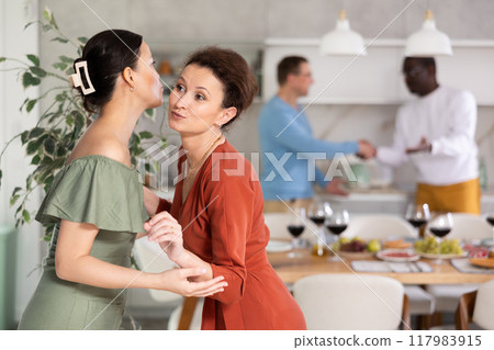 Women greet each other with kiss on cheek before festive dinner. Men shaking hands in background 117983915