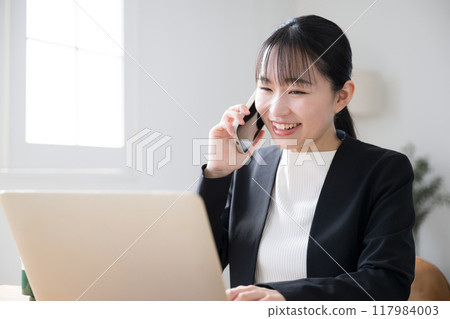 一位女商人一邊操作電腦一邊打電話，微笑著對著螢幕說話 117984003