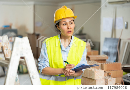 Female foreman in a protective helmet and vest checks the completed work on the repair of room Female foreman in a protective helmet and vest checks the completed work on the repair of room 117984028