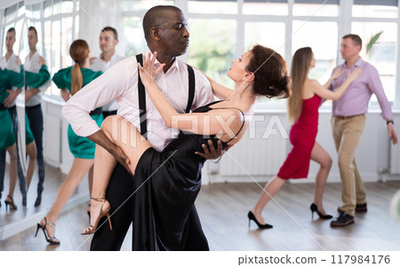 Elegant duet of man and woman in festive dresses dances passionate tango during group dance lesson in choreographic studio 117984176