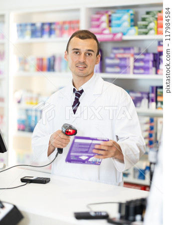 Man pharmacist in cash area works with computer and goods, scans drugs, checks availability Man pharmacist in cash area works with computer and goods, scans drugs, checks availability 117984584