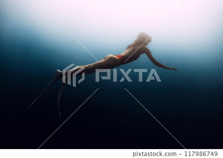 Gorgeous female freediver swims underwater in deep blue ocean. Sexy woman silhouette undersea. Gorgeous female freediver swims underwater in deep blue ocean. Sexy woman silhouette undersea. 117986749