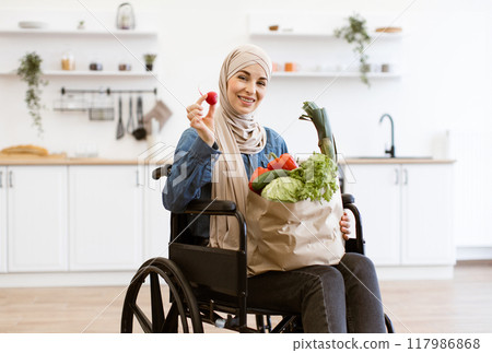 Muslim woman in wheelchair holding paper bag with vegetables in kitchen Muslim woman in wheelchair holding paper bag with vegetables in kitchen 117986868