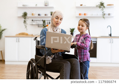 Woman in wheelchair working on laptop with young girl in kitchen Woman in wheelchair working on laptop with young girl in kitchen 117986874