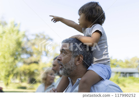 Curious little girl pointing at something while sitting on her grandfather's shoulders in the park, surrounded by family 117990252