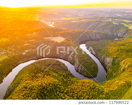 The sun sets dramatically over the Vltava River, casting a warm golden glow on the lush green hills and meandering waterways of Czechia, creating a picturesque landscape at dusk. 117990522