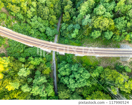 The Zampach stone railway bridge curves over lush greenery, showcasing a blend of nature and engineering in Czechia. The railway tracks extend across a vibrant forest landscape. 117990524