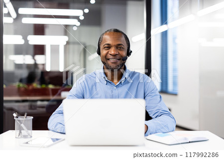 Smiling professional wearing headset sitting at desk with laptop, engaging in virtual meeting in bright office. Represents online communication, customer service, and remote work efficiency. 117992286