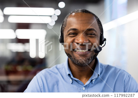 Mature African American businessman wearing headset inside modern office. Smiling confidently, embodying effective customer service and communication. Represents professionalism. 117992287