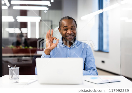 Confident businessman engages in a video call using laptop, showing ok gesture, highlighting positive communication. Reflects modern office environment 117992289
