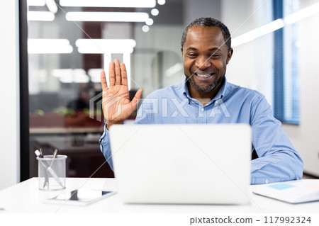 Smiling businessman in office environment engaged video call using laptop. Man waves hand at screen, showcasing friendly interaction during virtual meeting. Concept of remote work and communication. 117992324