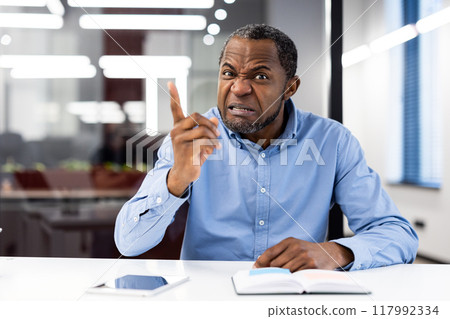 Frustrated businessman wearing blue shirt showing anger during video call, gesturing emphatically with finger. Intense expression during virtual meeting in modern office setting 117992334