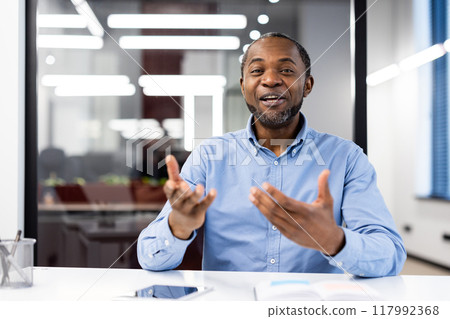 Confident professional man in blue shirt engaging in animated discussion during meeting in modern office. Expressive gestures add dynamism to conversation 117992368