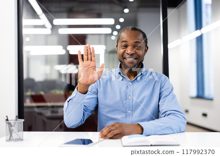 Smiling professional waves hand during video call in modern office. Engaging in virtual communication , creating friendly atmosphere. Represents business, communication, teamwork, technology 117992370