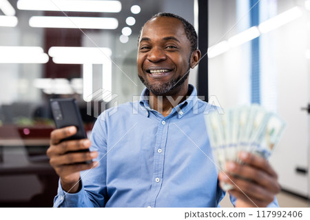 Smiling businessman holding stack of cash while using smartphone in office, showing financial achievement and joy. Image captures success, happiness, and technology in professional environment. 117992406