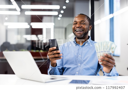 Confident African American businessman happily holding cash and phone in bright, modern office. Professional setting with focus on success and achievement. 117992407