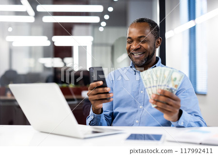 Smiling businessman sitting in modern office, holding cash and phone, symbolizing success and financial prosperity. Laptop on desk suggests productive work environment and technological engagement. Smiling businessman sitting in modern office, holding cash and phone, symbolizing success and financial prosperity. Laptop on desk suggests productive work environment and technological engagement. 117992411