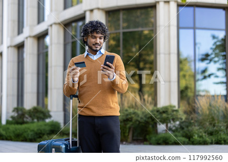 Confident man outside modern building using smartphone and holding credit card. Concept of online shopping, travel, and technology. Carrying suitcase, blending business urban environment. 117992560