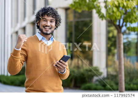 Businessman wearing casual attire smiling with joy while holding smartphone. Outdoor setting reflects professional success and positive emotions. Businessman wearing casual attire smiling with joy while holding smartphone. Outdoor setting reflects professional success and positive emotions. 117992574