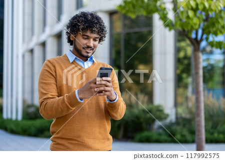 Young man enjoying outdoor work break, standing with phone in hand. Wearing casual sweater, he engages with technology in urban environment surrounded by nature, reflecting modern lifestyle Young man enjoying outdoor work break, standing with phone in hand. Wearing casual sweater, he engages with technology in urban environment surrounded by nature, reflecting modern lifestyle 117992575