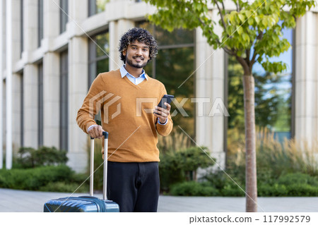 Smiling man stands outdoors with phone and suitcase, conveying readiness and confidence. He is dressed casually, suggesting a blend of business travel and leisure. 117992579