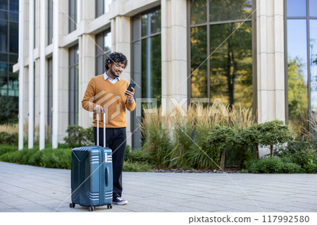 Smiling business traveler using smartphone while pulling suitcase in urban setting. Person wearing orange sweater walking outside office building. Captures concept of business travel Smiling business traveler using smartphone while pulling suitcase in urban setting. Person wearing orange sweater walking outside office building. Captures concept of business travel 117992580