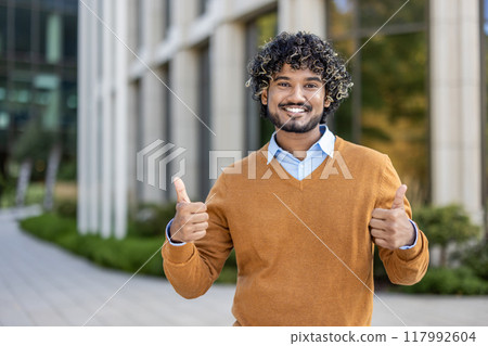 Young hispanic businessman standing outside office building expressing positivity with thumbs up gesture. He conveys confidence, success, and positive attitude in professional environment Young hispanic businessman standing outside office building expressing positivity with thumbs up gesture. He conveys confidence, success, and positive attitude in professional environment 117992604