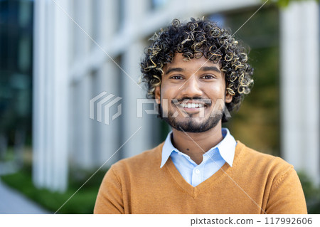 Confident young professional with curly hair and warm smile standing outdoors near modern office building. Image conveys success, positivity, and approachable demeanor in business context 117992606