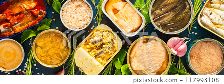 Enjoy an assortment of canned fish including salmon, mackerel, sprat, sardine, tuna, herring, and fish pate, displayed in open tins with spices and herbs on a blue background, banner Enjoy an assortment of canned fish including salmon, mackerel, sprat, sardine, tuna, herring, and fish pate, displayed in open tins with spices and herbs on a blue background, banner 117993108