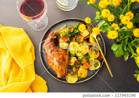 Baked rabbit with potatoes and carrots, a set table with food, a glass of wine and a vase of flowers Baked rabbit with potatoes and carrots, a set table with food, a glass of wine and a vase of flowers 117993190