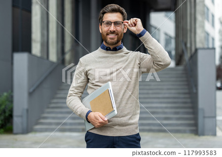 Portrait of mature teacher standing confidently in city environment. Holding notebook and adjusting glasses, he exudes professionalism and approachability, representing lifelong learning. 117993583