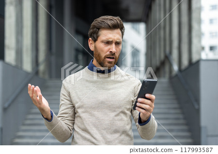 Businessman standing outside, looking frustrated at smartphone. Displaying expressions of confusion and stress. Depicts common challenges of modern work life. Businessman standing outside, looking frustrated at smartphone. Displaying expressions of confusion and stress. Depicts common challenges of modern work life. 117993587