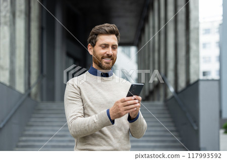 Confident man holding phone, smiling as he stands in front of corporate building. Represents business connectivity, technology, and communication. Ideal image for depicting professional lifestyle 117993592