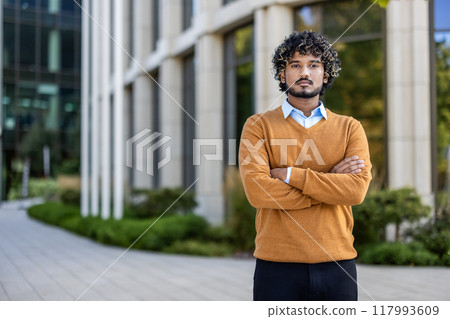 Confident hispanic businessman stands outside modern building with arms crossed, embodying success and determination. Image conveys professional confidence, leadership, and urban lifestyle. Confident hispanic businessman stands outside modern building with arms crossed, embodying success and determination. Image conveys professional confidence, leadership, and urban lifestyle. 117993609