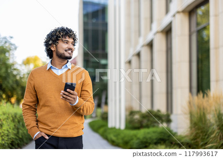 Hispanic businessman in orange sweater holding phone while walking outside office building. Smiling confidently and looking forward, showcasing modern urban work environment , professional lifestyle. Hispanic businessman in orange sweater holding phone while walking outside office building. Smiling confidently and looking forward, showcasing modern urban work environment , professional lifestyle. 117993617