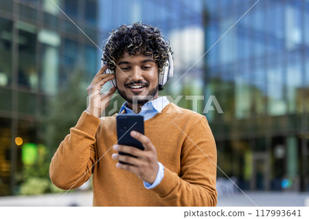 Smiling young man holds smartphone while wearing headphones in urban setting, enjoying music or communication. Dressed in autumn attire, displaying casual comfort outdoors. Captures modern lifestyle 117993641