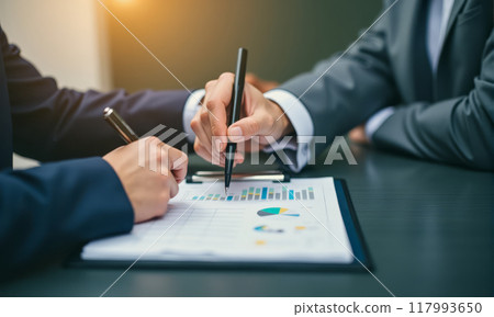 Professionals in business setting analyzing data charts, pen in hand pointing at paper, representing collaboration, decision-making, data analysis in corporate environment 117993650